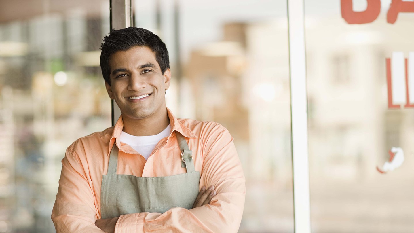 A young man, who is wearing an apron, is standing in front of the restaurant that he owns.